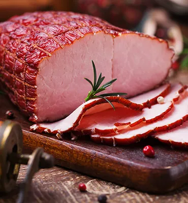 Sliced smoked ham on a wooden cutting board with sprigs of rosemary and scattered peppercorns.