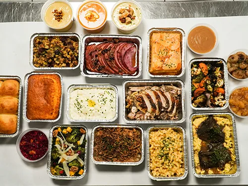 An assortment of various prepared dishes in foil containers, including meats, sides, bread, and desserts.