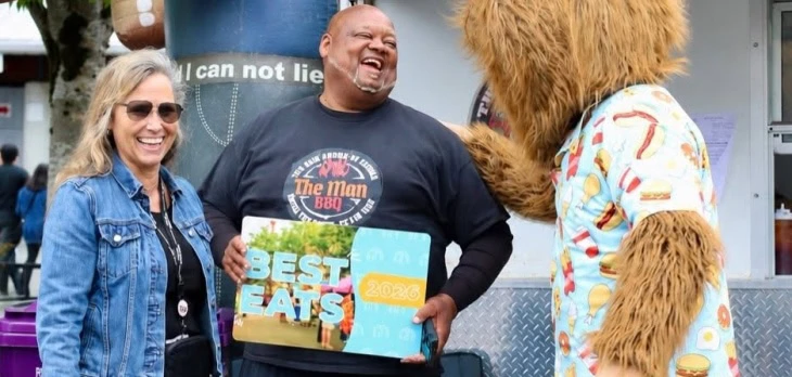 Three people, one in a bear costume, smile as a man holds a "Best Eats 2023" award sign at an outdoor event.